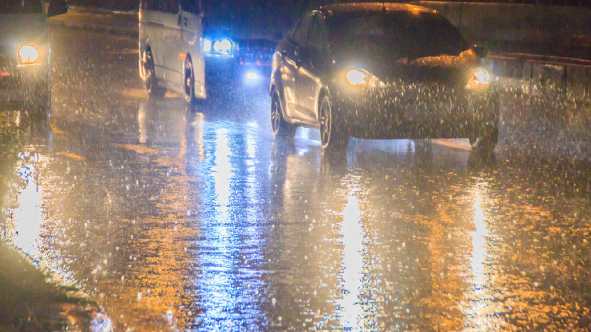 Several vehicles driving in the dark with headlights reflecting back from rain-soaked roads