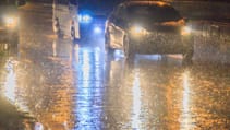 Several vehicles driving in the dark with headlights reflecting back from rain-soaked roads