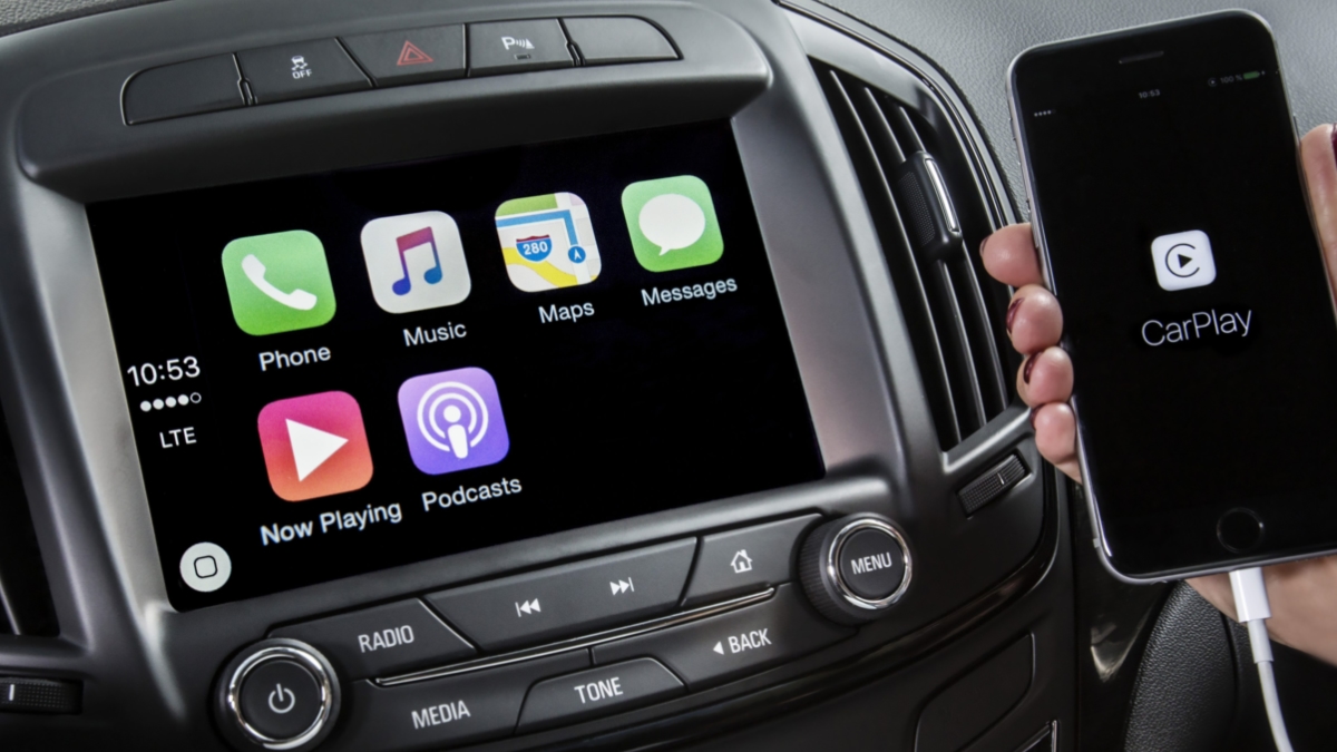 Woman holds mobile screen towards camera in front of centre console display, showing Apple CarPlay integration