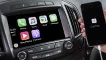 Woman holds mobile screen towards camera in front of centre console display, showing Apple CarPlay integration