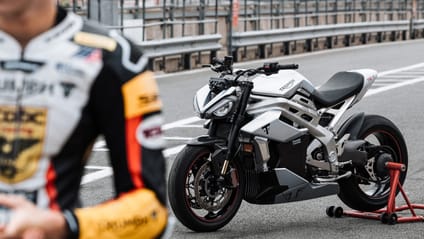 Triumph's prototype TE-1 sits mid-ground on the pit lane of a circuit, with the test rider in the foreground off to the left of the image