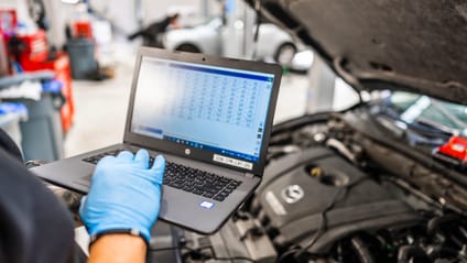 Garage mechanic looks at a computer interface with the bonnet of a used car up in the background