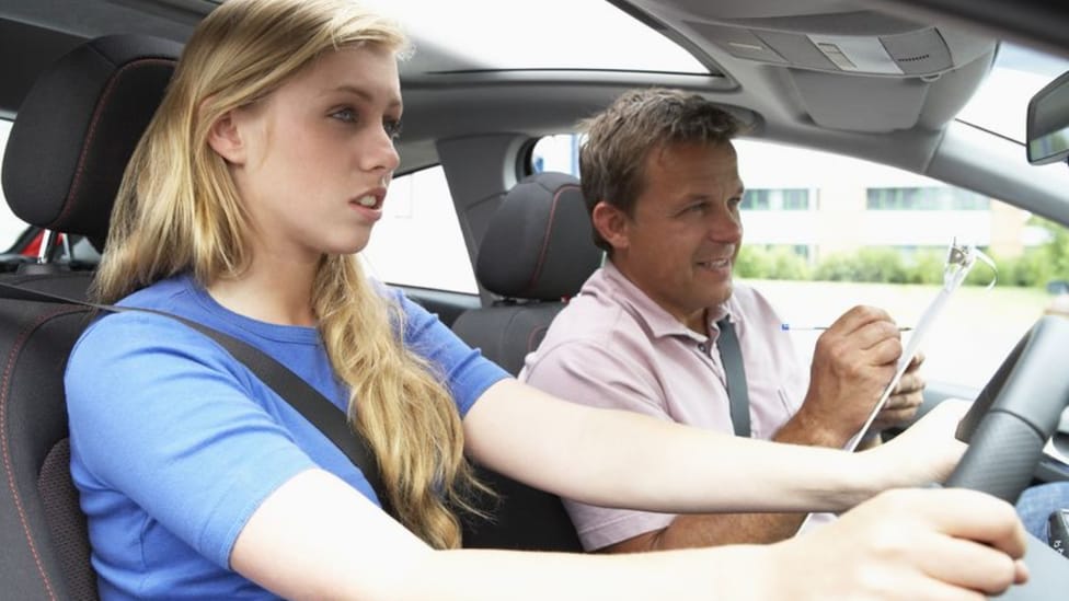 Male driving instructor gestures to female learner driver, in shot from driver's side window