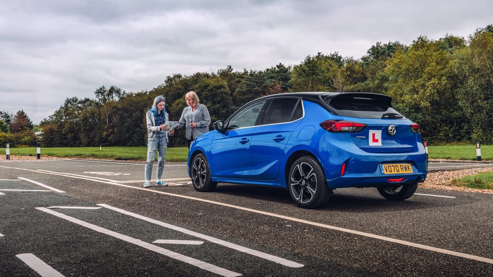 Female examiner stands next to female learner driver in front of learner car adorned with L plates