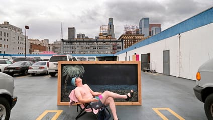 Man in front of a board with a beach drawn on it, wearing flip flops, in a car park