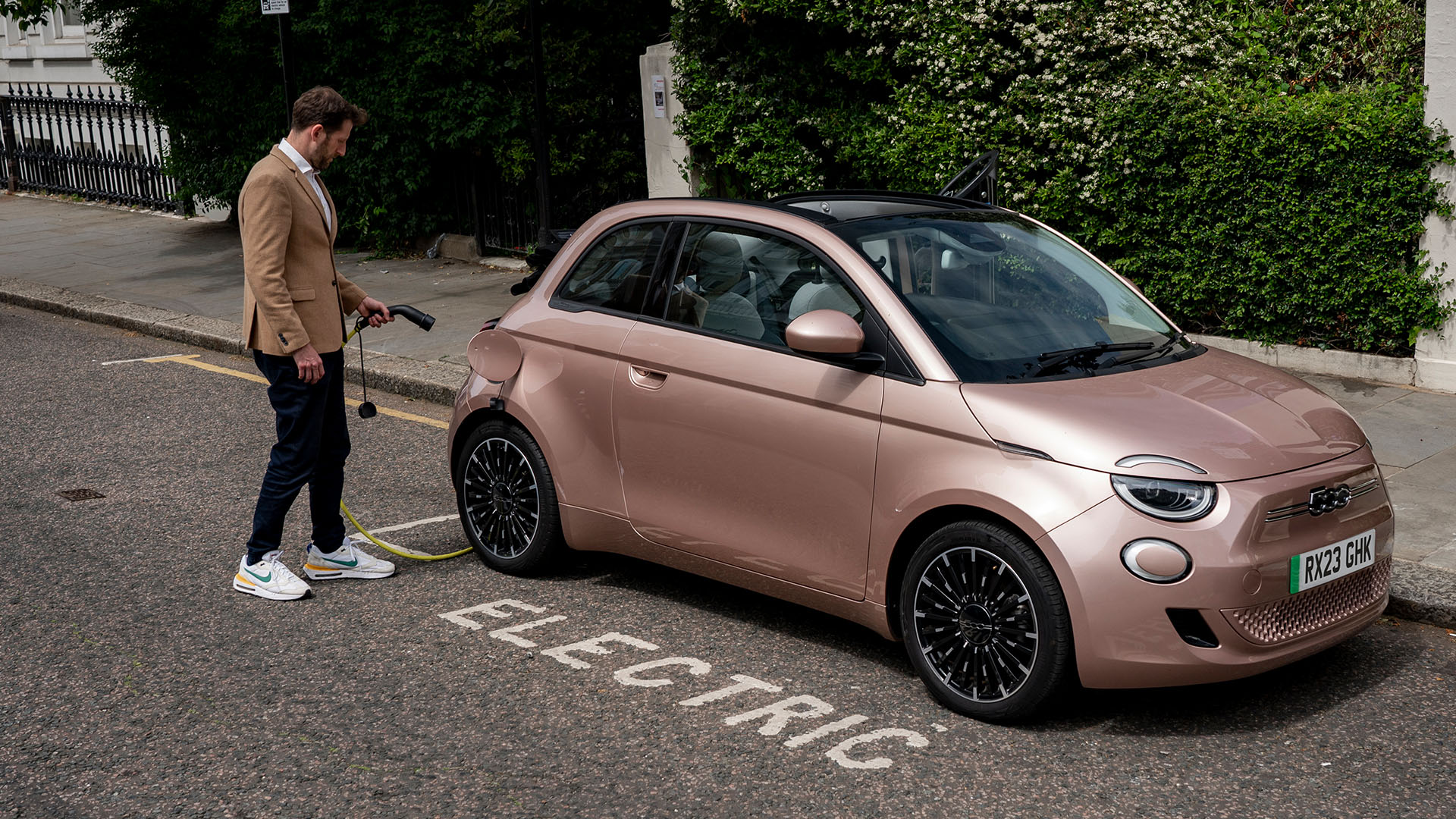 Guy approaching Fiat 500e on a leafy street, shot angled from front of car