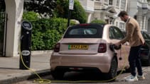 Guy plugging a Fiat 500e on a leafy street, shot angled from back of car