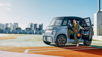 Static shot of Citroen Ami in car park with city backdrop. Girl getting out of car