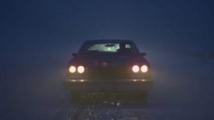 1977 Buick Riviera driving down a dark snowy road. Direct shot to camera head-on with headlights shining