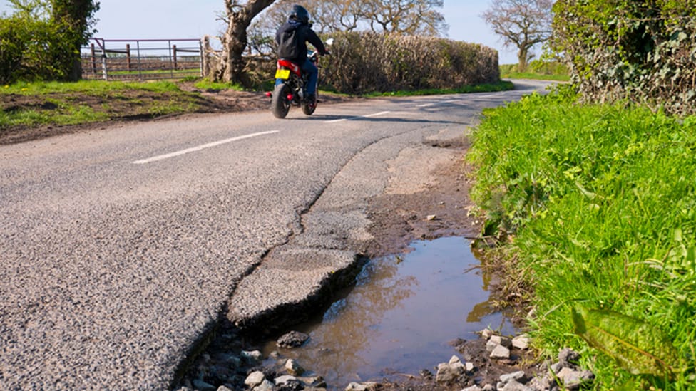 Close up of a pothole next to a green verge with a motorcyclist in the background