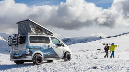 Electric camper parked on snowy mountain with two skiers walking away towards slope