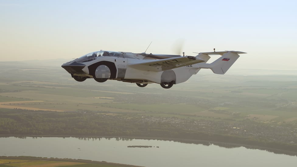 The AirCar airborne flying over a river with landscape in the background and the profile of the car with wings and tail clearly on show