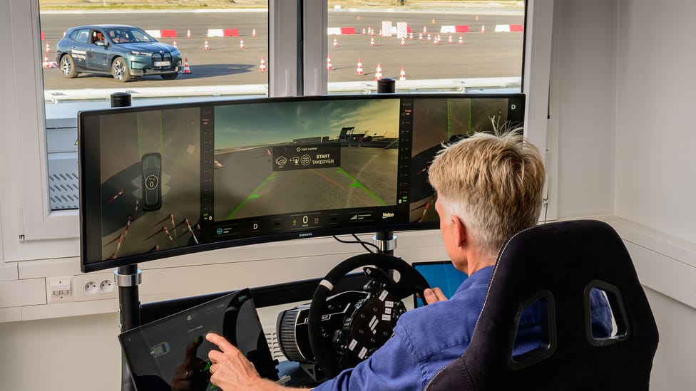 Man sitting at a self-driving module remotely driving a full-sized car which can be seen out on the tarmac through the window