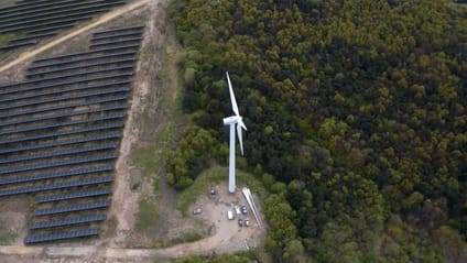 Bird's eye view of wind turbine with forest on right and solar farm on left