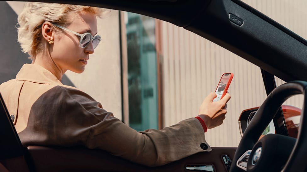 Woman holding iPhone in front of her while standing outside of a BMW model, shot taken from passenger side of vehicle through window