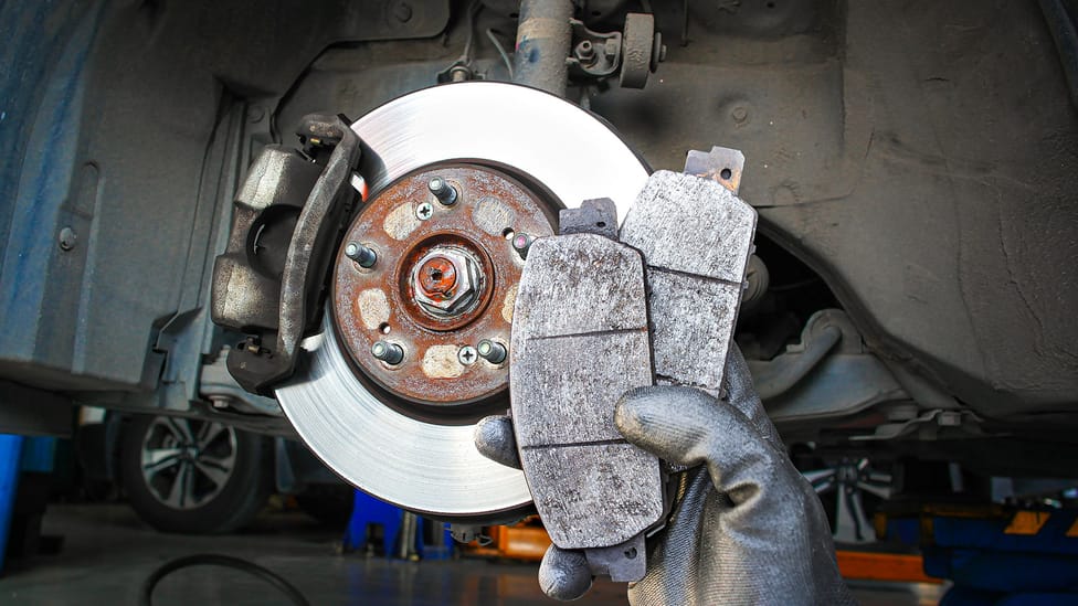 Mechanic's gloved hand holding up two worn brake pads in front of a brake disc