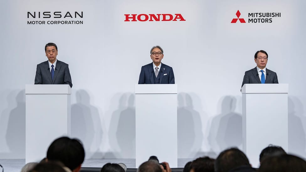 CEOs of Nissan, Mitusbishi and Honda standing at white lecturns with the audience in bottom third of image and respective company names above their heads