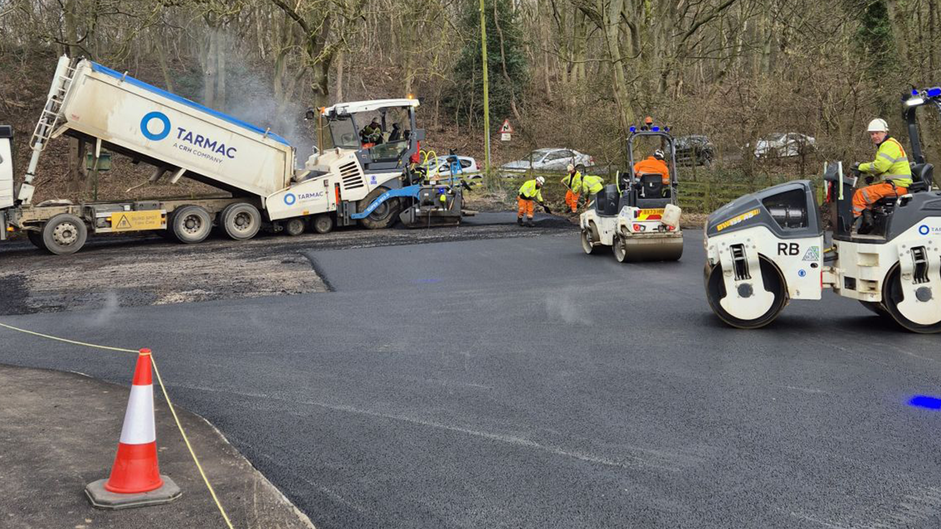 Wide shot of Tarmac dumper truck pouring new asphalt onto road with hi-vis jacketed workers in foreground