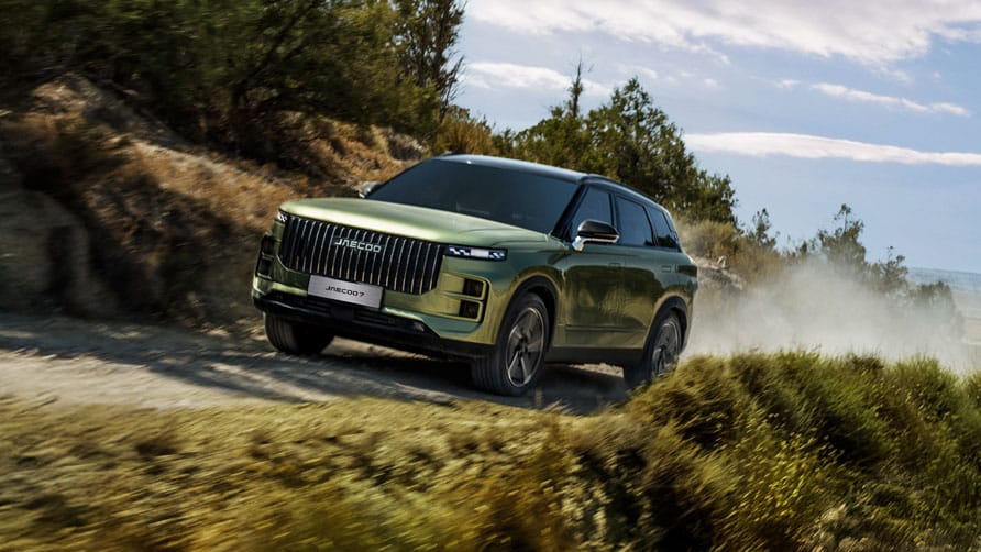 A sleek, green, hybrid JAECOO 7 SUV speeds down a dusty country road lined with trees. The sky is partly cloudy.