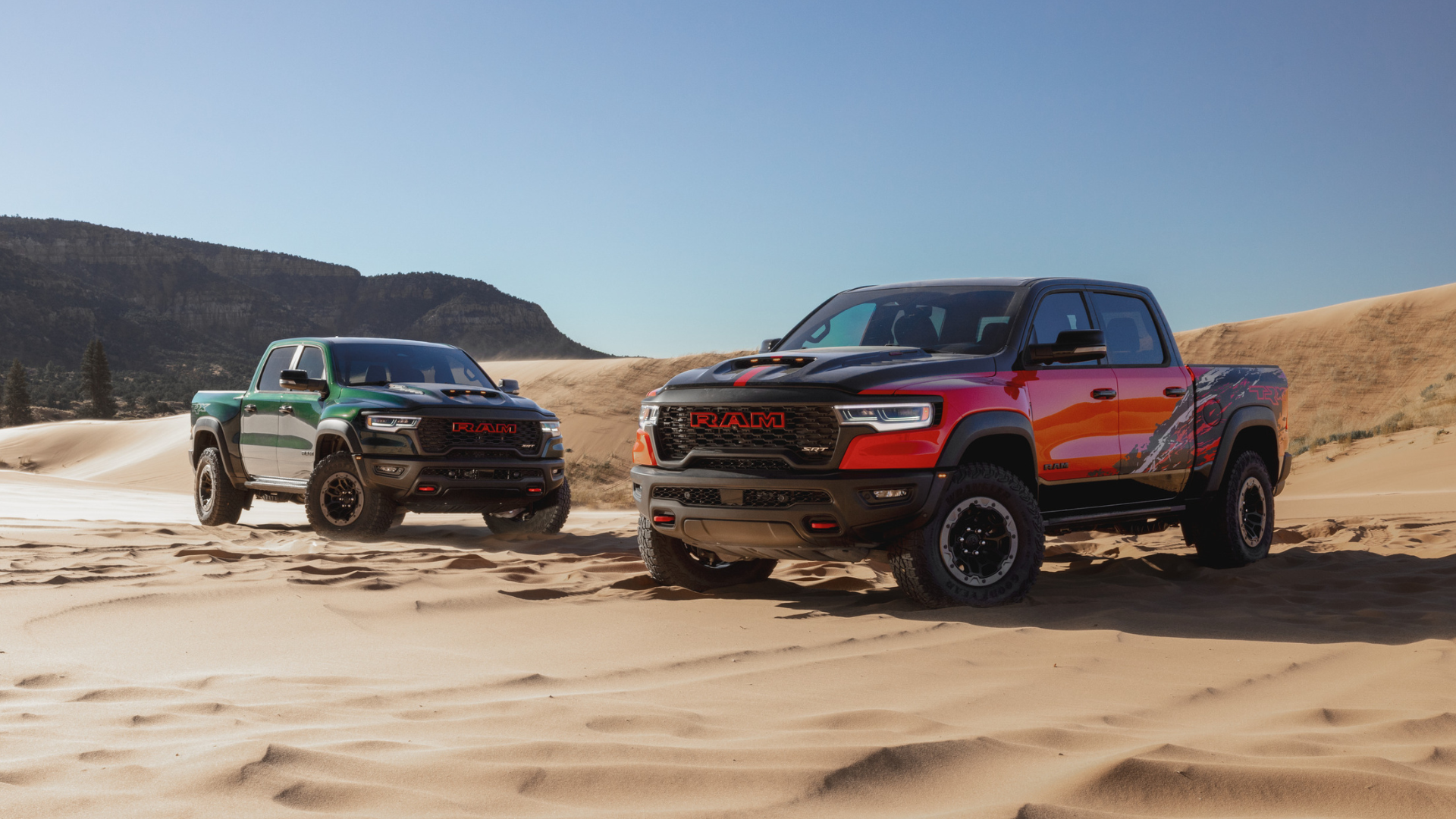 Two Ram 1500 SRT TRX's, one red and one green, are parked on a sand dune with mountains in the background.