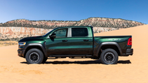 A green Ram TRX truck is parked on a sand dune against a backdrop of red rock formations under a blue sky.