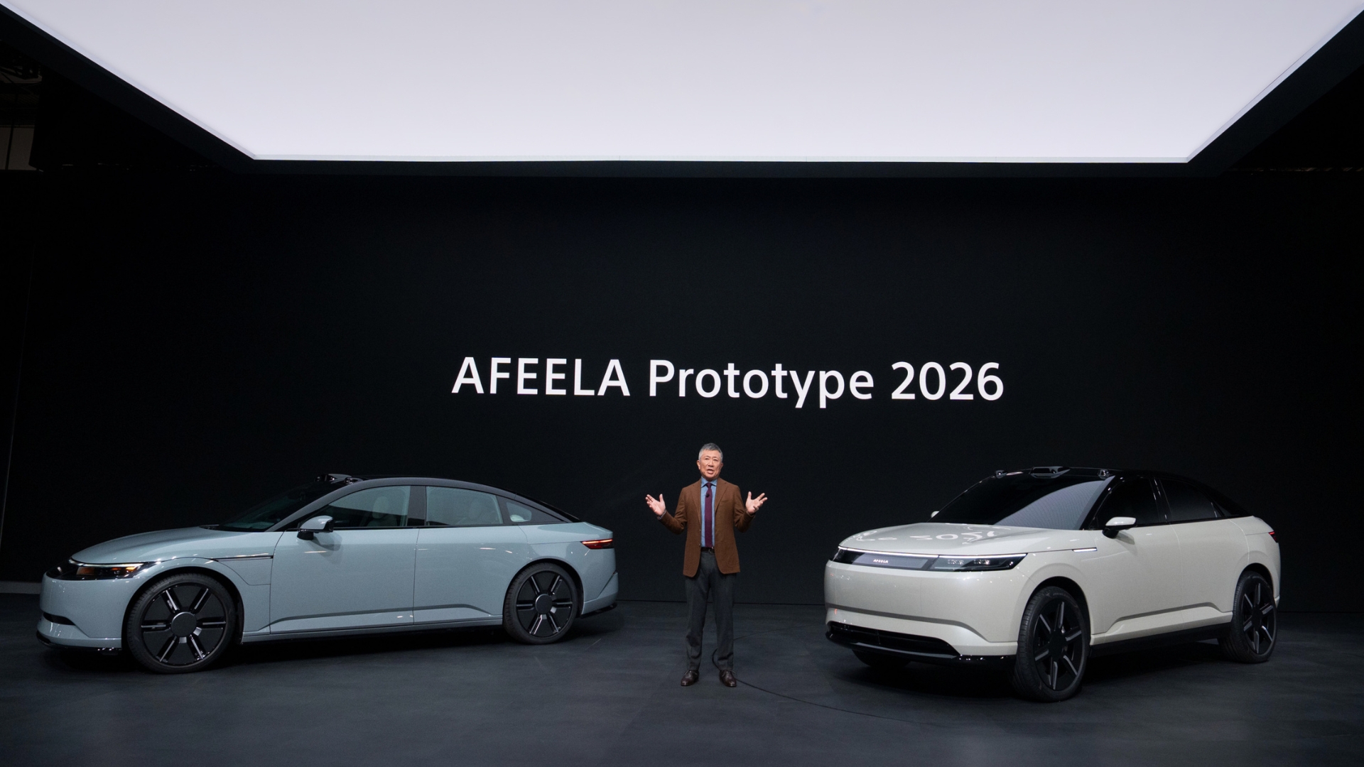 Two prototype Afeela 2026 cars, one light blue and one light beige, are displayed on a stage with a person standing between them.