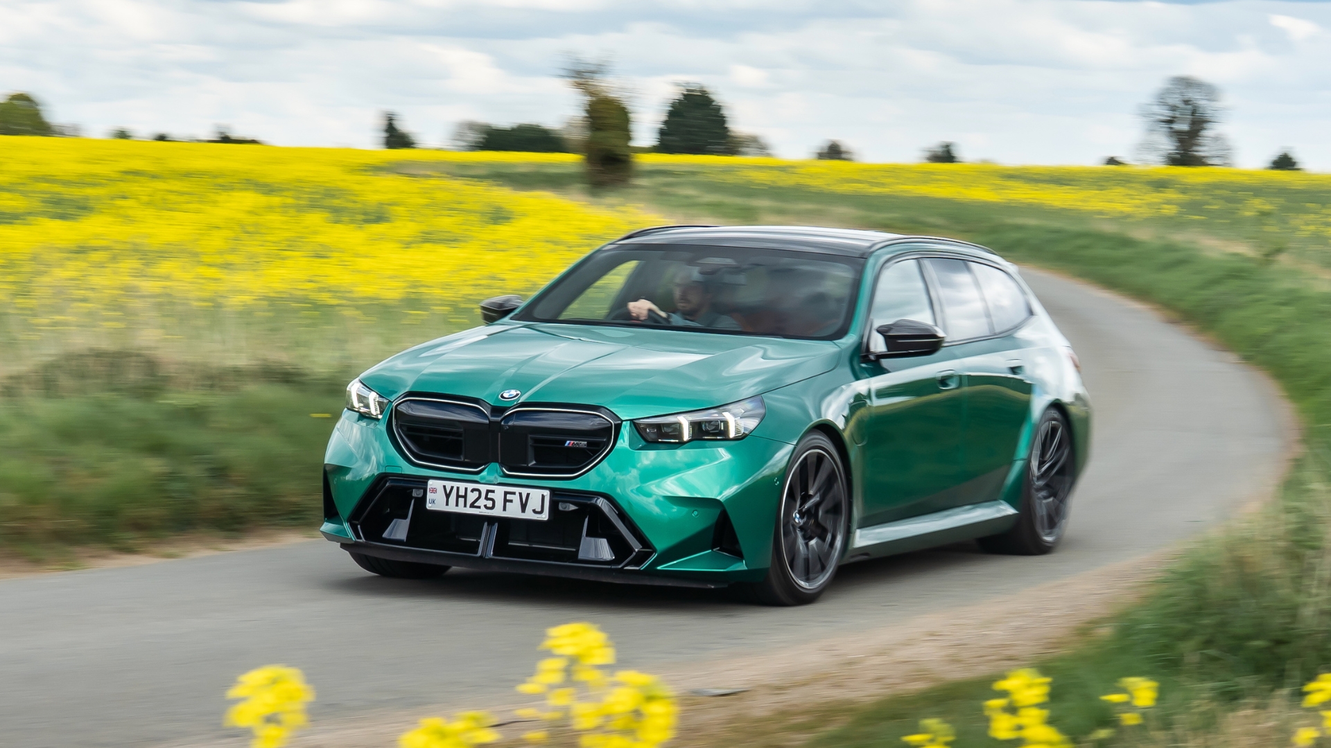 A green BMW drives on a road surrounded by a field of yellow flowers.
