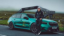 A person leans against a teal BMW M5 on a road, with a pedestrian bridge and foggy landscape in the background.