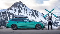 A green BMW with a roof cargo carrier is parked in front of a snowy mountain as someone holds up a pair of skis.