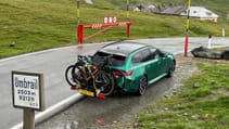 A green BMW with two bikes mounted on the back is parked next to a sign for Umbrail Pass in a mountainous area.
