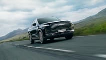 A sleek, black, hybrid JAECOO 7 SUV with a prominent grille cruises down an open road, surrounded by grassy hills and a cloudy sky