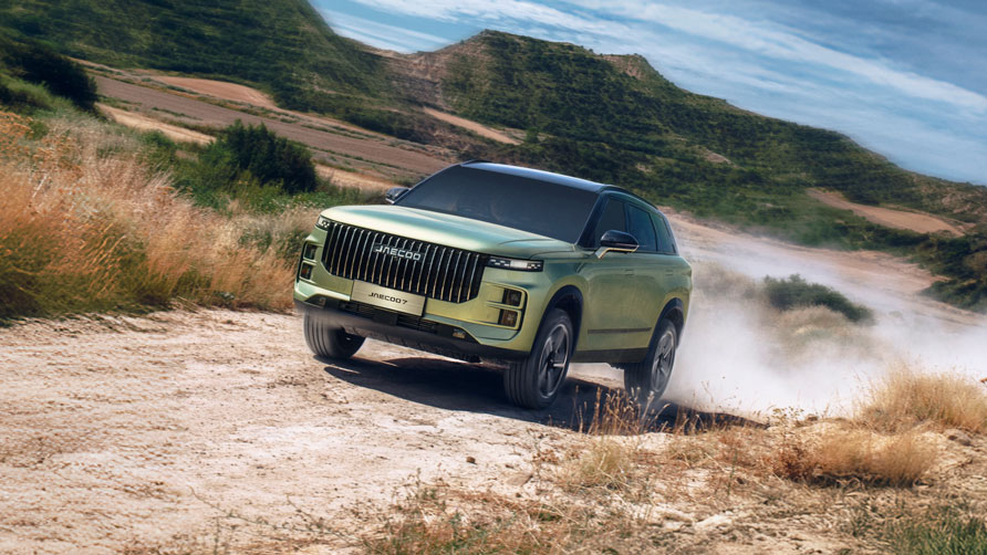 A sleek, green, hybrid JAECOO 7 SUV speeds down a dusty country road lined with trees. The sky is partly cloudy