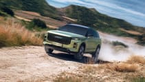 A sleek, green, hybrid JAECOO 7 SUV speeds down a dusty country road lined with trees. The sky is partly cloudy