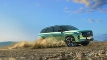 A sleek, green, hybrid JAECOO 7 SUV speeds down a dusty country road lined with trees. The sky is partly cloudy