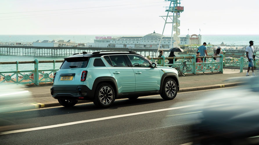 A pastel blue Renault 4 E-Tech electric car being driven on a seaside road in Brighton. The ocean and pier are visible in the background, while pedestrians enjoy the sunny day