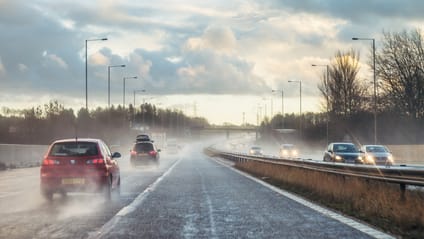 A motorway driving shot where several cars driving away from camera send up spray from the wet road, sun is trying to break through cloud